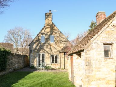 A stone cottage with a patio table and chairs and a small stone outbuilding in a garden at Cowfair Cottage in Chipping Campden