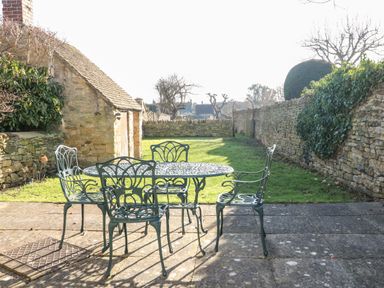 A stone patio with a metal table and four chairs next to a stone building and garden at Cowfair Cottage in Chipping Campden
