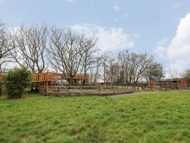 An outdoor playground with a wooden play structure slide fenced area and sheds in a grassy field at Tynewydd Fields in Cross Inn