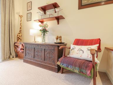 A wooden carved chest with a lamp and flowers on top next to a chair with a cushion and throws in a room at Aelwyd Y Don in Tresaith