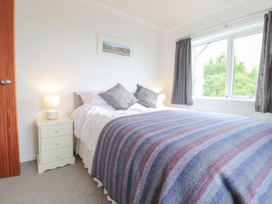 A bedroom with a bed covered by a striped blanket and two pillows next to a nightstand with a lamp and a window with curtains at Aelwyd Y Don in Tresaith