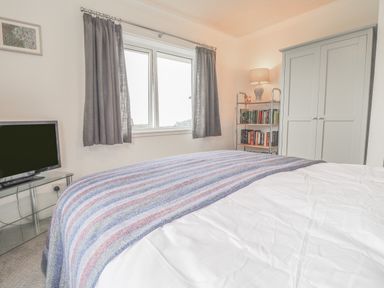 A bedroom with a bed covered by a striped blanket a wardrobe a bookshelf with a lamp a window with curtains and a television on a glass stand at Aelwyd Y Don in Tresaith