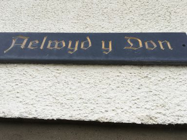 A black rectangular sign with gold Gothic lettering on a white textured wall at Aelwyd Y Don in Tresaith