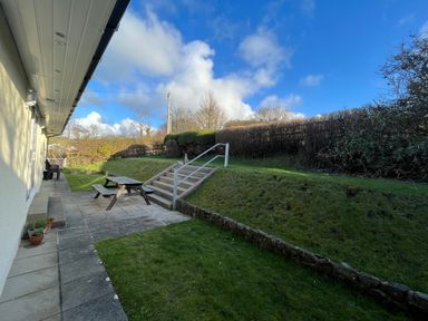 An outdoor patio area with a wooden picnic table and benches next to a building with steps leading up to a grassy area and hedges at Aelwyd Y Don in Tresaith