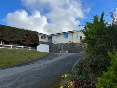 A driveway leading up to a house with stone walls and garage with bushes and flowers in the foreground at Aelwyd Y Don in Tresaith