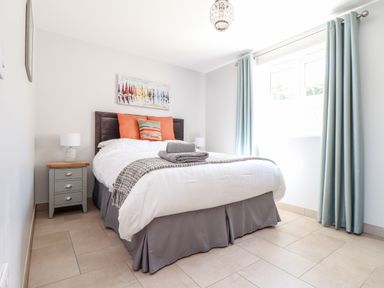 A bedroom with a double bed pillows bedside table lamp window with curtains and painting on wall at Chy-Whel in Sticker