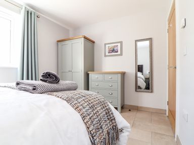 A bedroom with a bed folded blankets a wardrobe a chest of drawers a mirror a painting curtains and a wooden door at Chy-Whel in Sticker