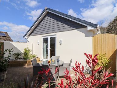An outdoor patio area with a round glass table and four chairs next to a garden bed and a house with double glass doors at Chy-Whel in Sticker