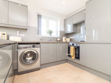 A kitchen with grey cabinets a front loading washing machine an oven with a yellow towel on the handle and a window with a plant on the sill at Chy-Whel in Sticker
