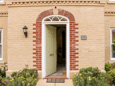 The front entrance of a brick house with a partially open pale green door and yellow flowers by the path at Coral House in Overstrand