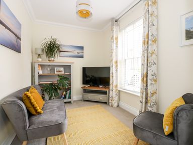 A small living room with two gray chairs with yellow pillows a bookshelf a television on a stand and a window with floral curtains at Coral House in Overstrand