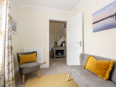 A room with a gray armchair and sofa with yellow cushions a yellow patterned rug floral curtains a white door and table in a hallway at Coral House in Overstrand
