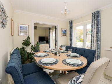 A dining area with a round wooden table set with plates and wine glasses surrounded by blue and beige upholstered chairs with curtains and wall art at Coral House in Overstrand