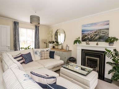 A living room with striped sofas surrounded by cushions a fireplace with plants a cabinet with a round mirror and window with curtains at Coral House in Overstrand