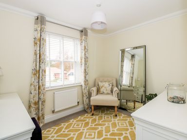 A small room with an armchair a large mirror a window with curtains a rug and white dressers at Coral House in Overstrand