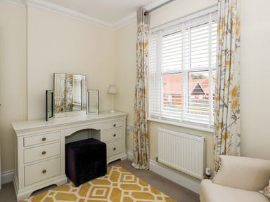 A bedroom corner with a white vanity table with a mirror and a lamp a black ottoman a window with white blinds and floral curtains a radiator and a beige armchair at Coral House in Overstrand