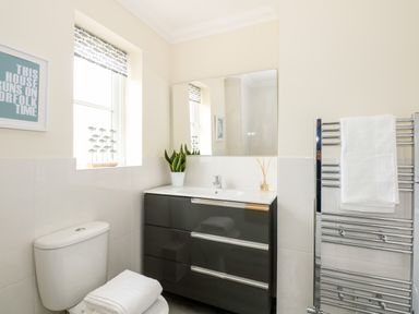 A bathroom with a toilet and folded towels a sink with a cabinet and a plant a window with blinds and a towel rack at Coral House in Overstrand