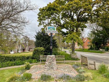 A village sign for Overstrand surrounded by benches trees and houses along a road in a grassy area at Coral House in Overstrand