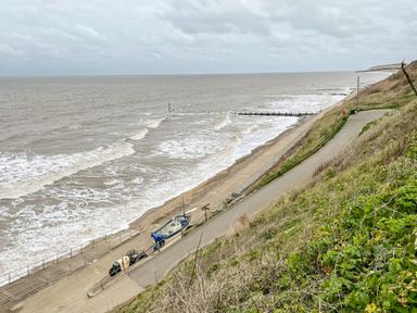 A coast with waves hitting the shore a pathway running parallel to the beach and a boat near the shore at Coral House in Overstrand