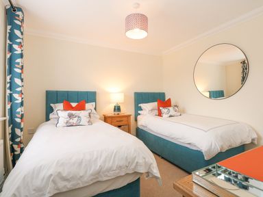 A bedroom with two single beds a wooden nightstand with a lamp a round wall mirror and patterned curtains at Coral House in Overstrand