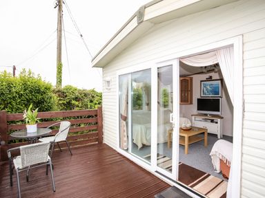 A patio with two chairs and a small table next to a cabin with sliding glass doors revealing a living area with a sofa a coffee table and a television inside at Bwthyn Y Bae Lodge Church Bay near Llanfaethlu
