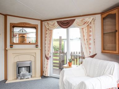 A living room with a fireplace a white covered sofa glass cabinet and glass door with curtains at Bwthyn Y Bae Lodge in Church Bay near Llanfaethlu