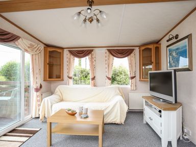 A living room with a sofa covered in a light throw a wooden coffee table a television on a white stand and curtains on windows at Bwthyn Y Bae Lodge in Church Bay near Llanfaethlu