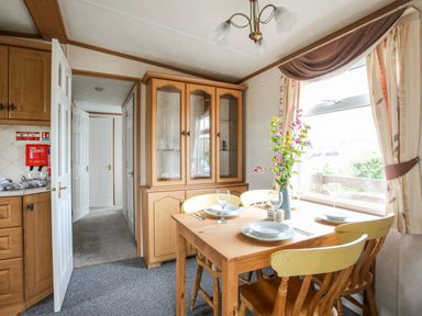 A dining area with a wooden table set with plates glasses and flowers by a window and a glass cabinet in the background at Bwthyn Y Bae Lodge in Church Bay near Llanfaethlu