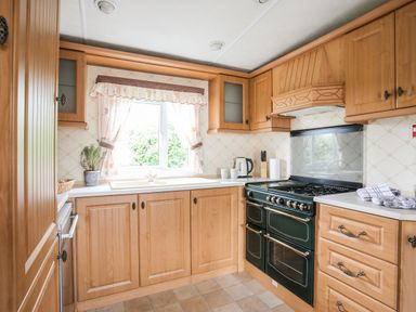 A kitchen with wooden cabinets a window with curtains a sink a stove a kettle and some mugs at Bwthyn Y Bae Lodge in Church Bay near Llanfaethlu