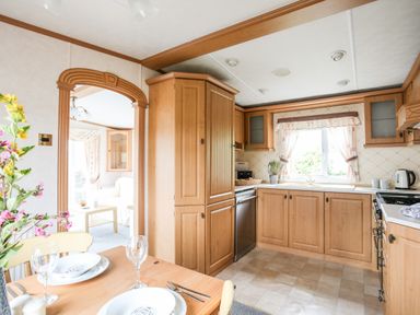 A kitchen with wooden cabinets a table set with plates and glasses and a view into a living room at Bwthyn Y Bae Lodge in Church Bay near Llanfaethlu