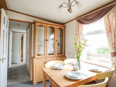 A dining area with a wooden table set with plates glasses and cutlery next to a window with curtains and a wooden cabinet against the wall at Bwthyn Y Bae Lodge in Church Bay near Llanfaethlu