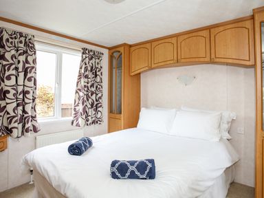 A bedroom with a double bed white bedding blue towels wooden cabinets and patterned curtains at Bwthyn Y Bae Lodge in Church Bay near Llanfaethlu