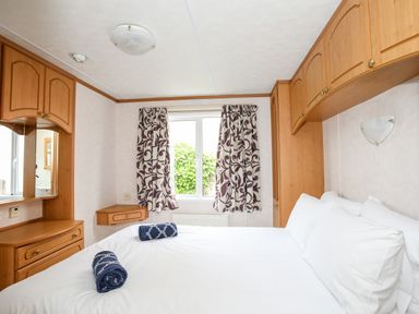 A bedroom with a white bed two blue towels wooden cabinets and a window with patterned curtains at Bwthyn Y Bae Lodge in Church Bay near Llanfaethlu
