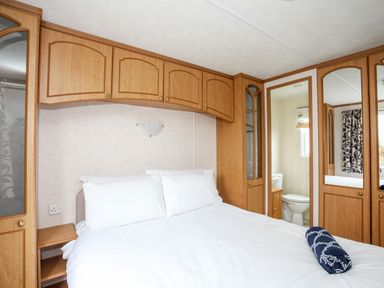 A bedroom with a bed white pillows wooden cabinets and a bathroom visible through a doorway at Bwthyn Y Bae Lodge in Church Bay near Llanfaethlu