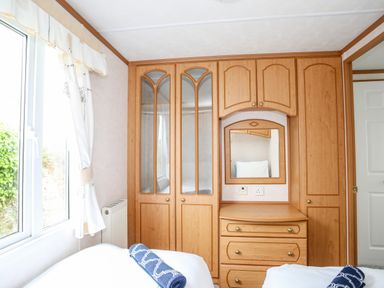 A bedroom with wooden wardrobes a mirror and a window with curtains at Bwthyn Y Bae Lodge in Church Bay near Llanfaethlu