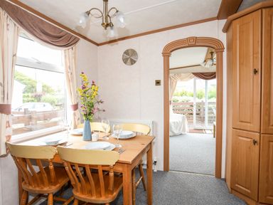 A dining area with a wooden table set for four with a vase of flowers next to a large window at Bwthyn Y Bae Lodge in Church Bay near Llanfaethlu