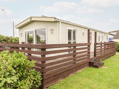 A single story mobile home with beige siding and a brown wooden deck surrounded by green grass and plants at Bwthyn Y Bae Lodge in Church Bay near Llanfaethlu