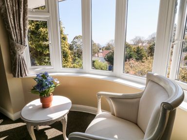 A white chair and small table with a potted plant by bay windows showing trees and houses at Harbour Lights in Torquay