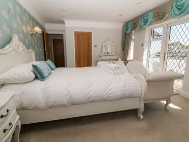 A bedroom with a bed and white bedding two blue pillows a white chair a dresser with a mirror and windows with blue curtains at Harbour Lights in Torquay