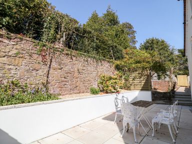 An outdoor patio with a white metal table and chairs beside a stone wall and garden at Harbour Lights in Torquay