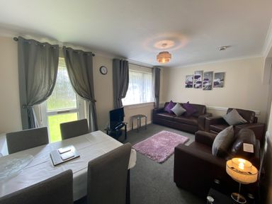 A living room with brown sofas and purple cushions a dining table with chairs a small tv and windows with curtains at Lands End in Liskeard near St Cleer