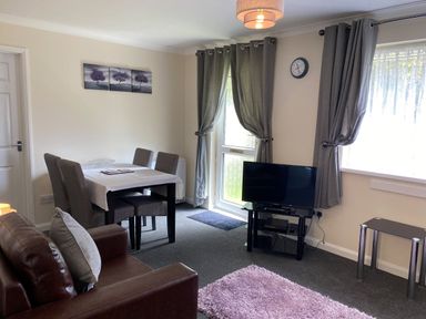 A living room with a dining table and chairs a television on a stand and a brown sofa at Lands End in Liskeard near St Cleer