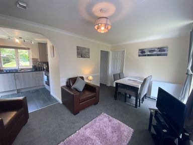 A living room with two brown armchairs a dining table with four chairs a television and a view into a kitchen at Lands End in Liskeard near St Cleer