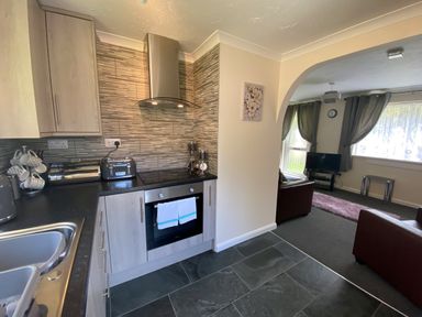 A kitchen with cabinets a toaster an oven and a sink next to a living room with a television and armchairs at Lands End in Liskeard near St Cleer