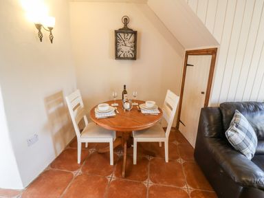 A dining area with a round wooden table set for two with white chairs a wall clock and a leather sofa at Bwthyn Siop Pencaerau in Rhiw near Aberdaron