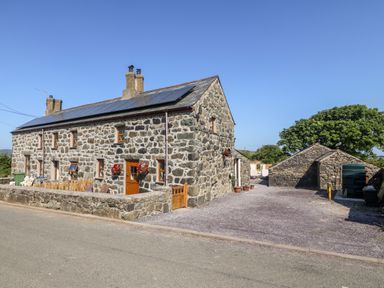 A stone house with wooden doors and windows next to a gravel driveway and outbuildings at Bwthyn Siop Pencaerau in Rhiw near Aberdaron