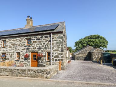 A stone cottage with wooden doors and windows and flower baskets outside with adjacent stone buildings and a tree at Bwthyn Siop Pencaerau in Rhiw near Aberdaron