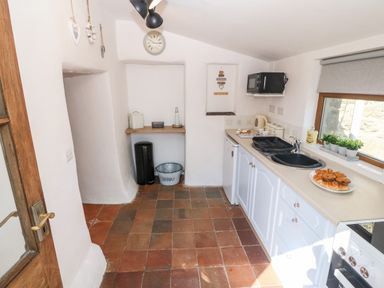 A kitchen with tiled floor white cabinets a sink a microwave a kettle and pastries on the counter at Bwthyn Siop Pencaerau in Rhiw near Aberdaron