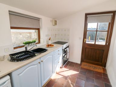 A kitchen with a sink, dish rack, stove, wooden door with a welcome mat, and window with grey blinds at Bwthyn Siop Pencaerau in Rhiw near Aberdaron