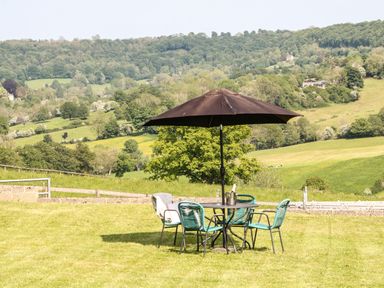 An outdoor table with four chairs and a large umbrella on grass with hills and trees in the background at Farm View Lodge in Painswick